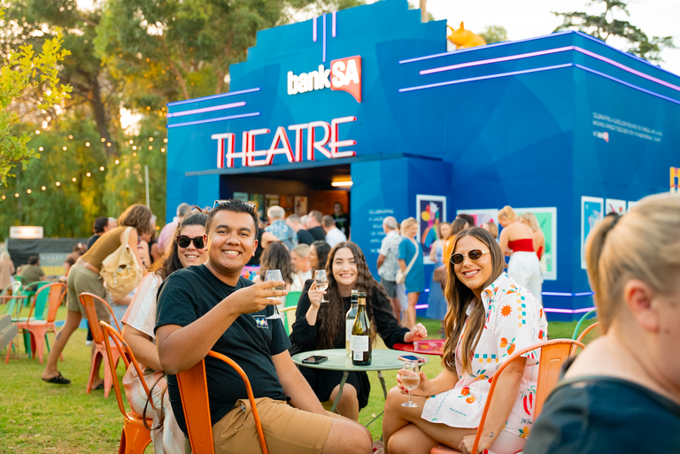 People enjoying drinks outdoors at the Adelaide Fringe Festival, sitting outside the blue BankSA Theatre venue on a sunny day in the park.